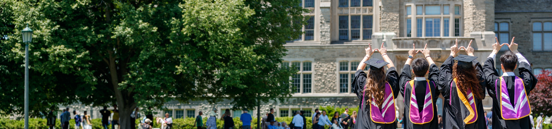 Four students in their graduation gowns and regalia's with their backs turned, holding their hands up making a W (for Western), facing UC tower.