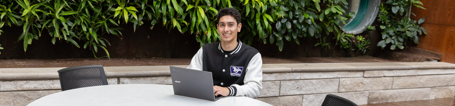 A student sitting at a table, typing on their laptop, wearing a Western jacket and smiling at the camera.