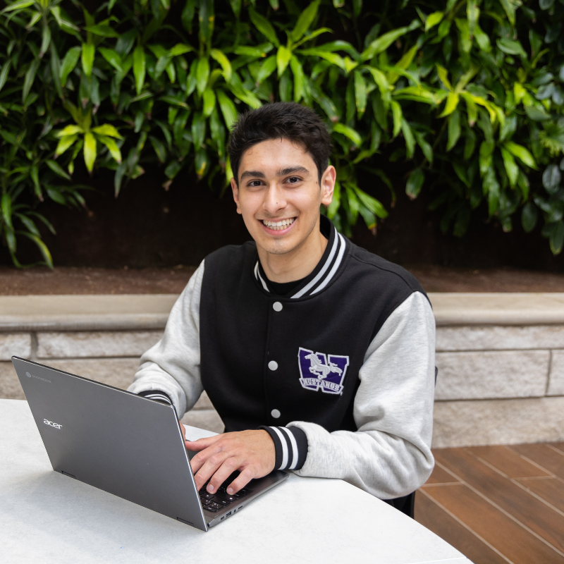 A student sitting at a table, typing on their laptop, wearing a Western jacket and smiling at the camera.
